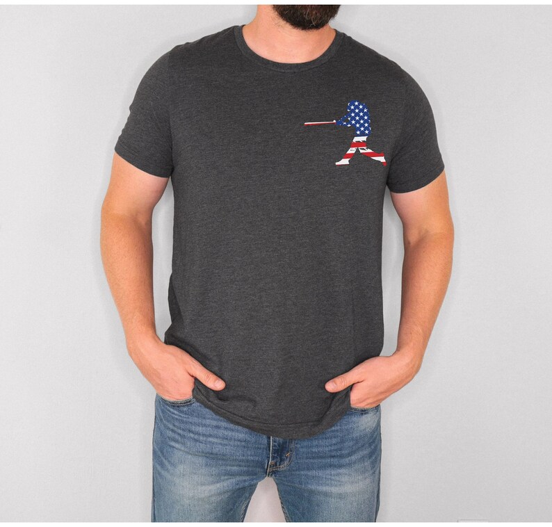 May include: A dark grey t-shirt with a baseball player silhouette printed on the left chest. The player is silhouetted in the colours of the American flag.
