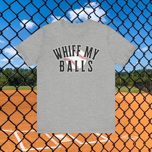 Puede incluir: Camiseta gris jaspeado con el texto "WHIFF MY BALLS" en blanco y negro. El gráfico de béisbol es rojo y blanco. El fondo es un campo de béisbol.
