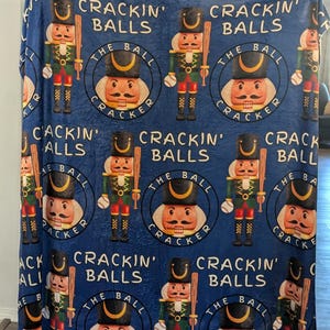 May include: A blue fleece blanket with a repeating pattern of nutcracker figures holding baseball bats. Each nutcracker is surrounded by a circle with the text "The Ball Cracker" and the phrase "Crackin' Balls".