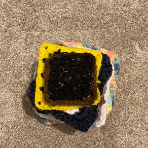 May include: A stack of crocheted squares in various colors, including yellow, brown, blue, white, and pink. The top square is yellow with a brown border and a black center.