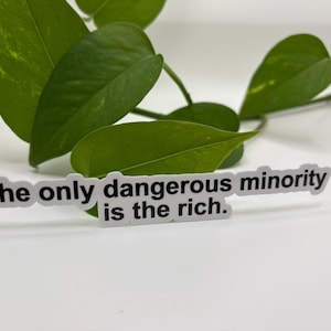 May include: A white sticker with black text that reads "The only dangerous minority is the rich." The sticker is on a white surface with green leaves in the background.
