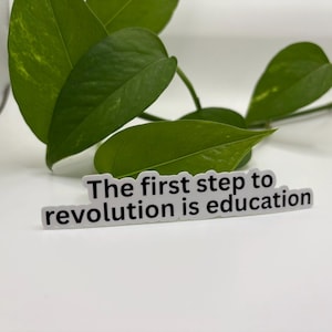 May include: A white sticker with the text "The first step to revolution is education" in black. The sticker is placed in front of green leaves.