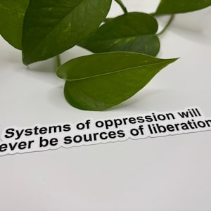 May include: A white sticker with black text that reads "Systems of oppression will never be sources of liberation." The sticker is on a white surface, with green leaves in the background.