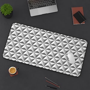 May include: A rectangular desk mat featuring a repeating geometric pattern of white, light gray, and dark gray triangles that create a three-dimensional effect. The mat has a smooth surface and a black border, with a white computer mouse placed on top.