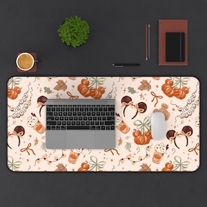 May include: A rectangular desk mat with a light cream background featuring a repeating pattern of orange pumpkins, brown mouse ears, coffee cups with whipped cream, bows, and garlands with the text 'TIS THE SEASON' in white circular beads.