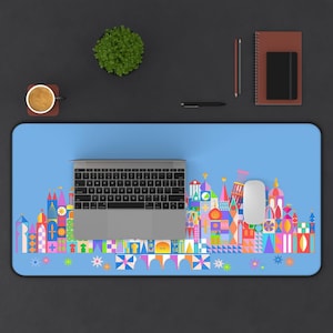 May include: A rectangular desk mat with a light blue background featuring a colorful, geometric skyline illustration of stylized buildings and towers in shades of pink, orange, yellow, green, and purple, with a laptop and mouse placed on top.