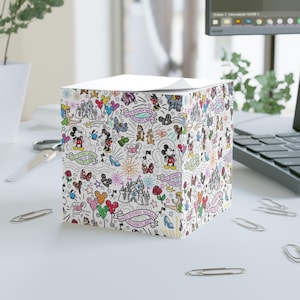May include: A white cube-shaped notepad dispenser with a colorful Disney-themed doodle pattern. The design includes Mickey and Minnie Mouse, castles, and the words "Walt Disney World" on banners. The notepad is on a white desk with paperclips.