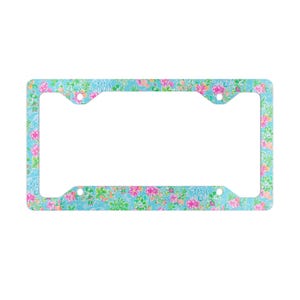 May include: A license plate frame with a floral pattern in shades of pink, green, and orange on a light blue background. The frame has a rectangular shape with rounded corners and mounting holes.