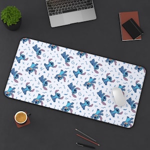 Op de afbeelding: Een rechthoekige bureaumat met een herhaald patroon van een blauw stripfiguur op een witte achtergrond. Het ontwerp bevat blauwe zigzags, cijfers en letters. Een computermuis ligt op de mat, met een laptop en andere kantoorbenodigdheden zichtbaar.