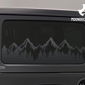 May include: Black Jeep Wrangler with a mountain range decal on the rear window. The decal is a silhouette of mountains and trees in a dark gray color. The decal is on the driver's side rear window. The decal says "MOONDECALSTORE" in white text.
