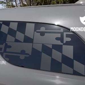 May include: A gray SUV with a black and white Maryland state flag decal on the rear window. The decal is a silhouette of the flag with the words "MOONDECALSTORE" printed below it.