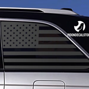 May include: A black car with a gray and black American flag decal on the back window. The decal is a silhouette of the American flag with the stars and stripes in black. The decal is on the back window of a car. The decal has the text "MOONDECALSTORE" on the bottom right corner.