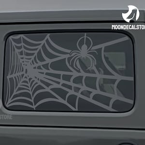 May include: A gray car with a spider web and spider decal on the back window. The decal is white and features a spider web with a spider in the center. The text "MOONDECALSTORE" is visible on the car.