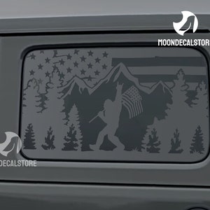May include: A dark gray decal featuring a silhouette of Bigfoot holding an American flag in a mountain and forest scene. The top of the decal has a flag design with stars and stripes. The decal is designed to be applied to a vehicle window.