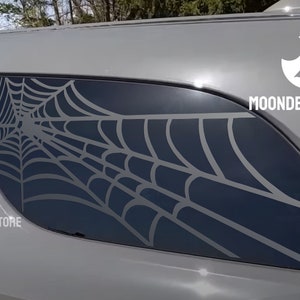May include: A gray car with a spiderweb decal on the back window. The decal is black and has a detailed design. The car has the text "MOONDECALSTORE" on the side.