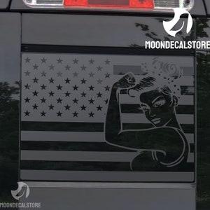 May include: A black and white decal of a woman with her arm raised in a 'we can do it' pose, superimposed on an American flag. The decal is on a black truck window.