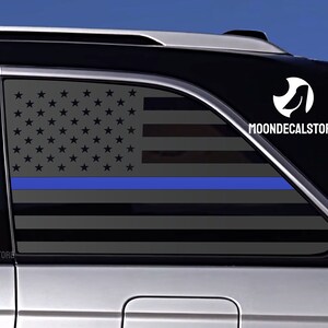 May include: A black car with a gray and blue American flag decal on the back window. The decal includes a thin blue line across the flag. The decal has the text "MOONDECALSTORE" on the right side.