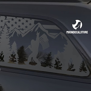 May include: A white vinyl decal of a Bigfoot holding an American flag in front of a mountain range and forest. The decal is applied to the back window of a black SUV. The decal includes the text "MOONDECALSTORE".