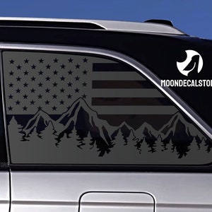 May include: A black and white decal of an American flag with mountains and trees in the foreground. The decal is applied to the back window of a silver car. The decal includes the text "MOONDECALSTORE".