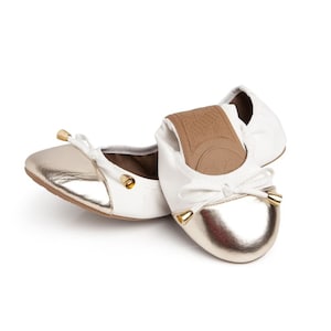 May include: A pair of white ballet flats with gold accents. The flats have a bow on the toe and a gold trim around the edge.