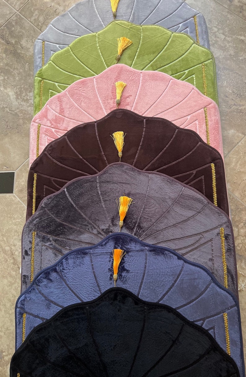 May include: Six prayer rugs in different colors, stacked on top of each other. The rugs are all the same shape, with a scalloped edge and a tassel at the top. The colors of the rugs are gray, green, pink, brown, purple, blue, and black.