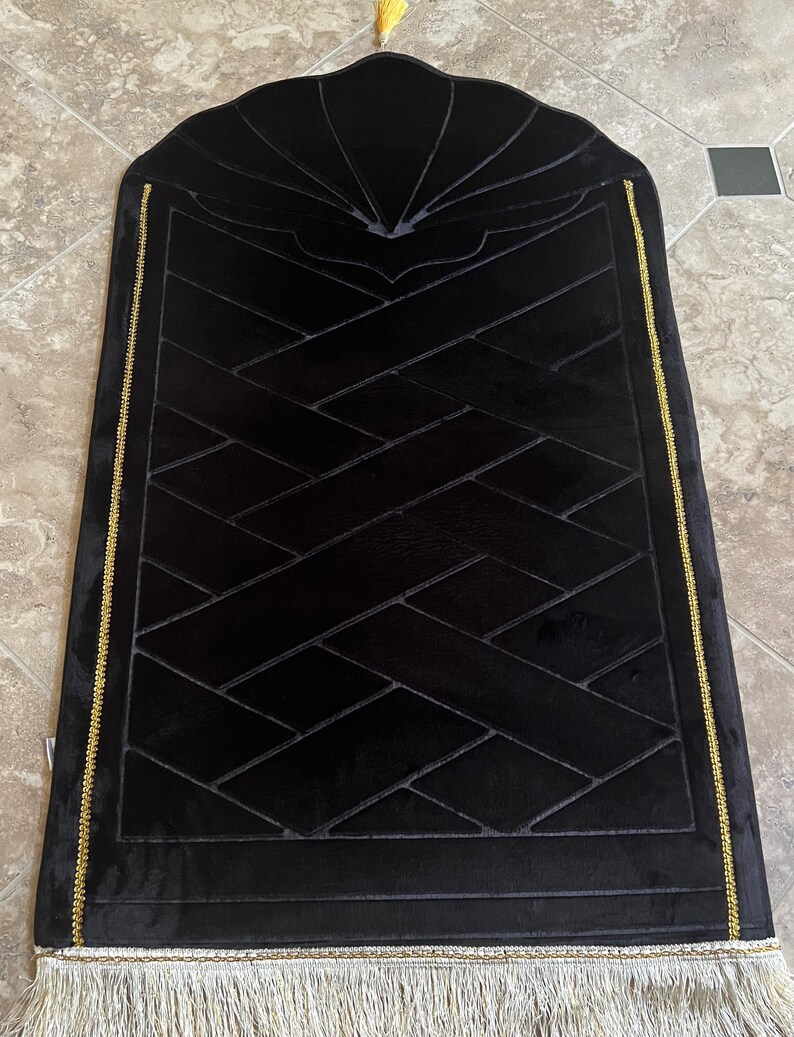 May include: Black prayer rug with a geometric pattern and gold trim. The rug has a tassel at the top and a fringe at the bottom.