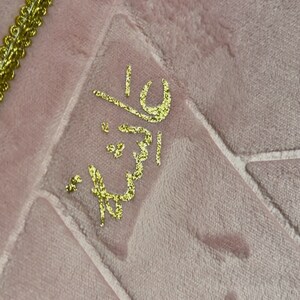 May include: A pink prayer rug with a gold embroidered design. The design features the Arabic word "Allah" in a decorative script.