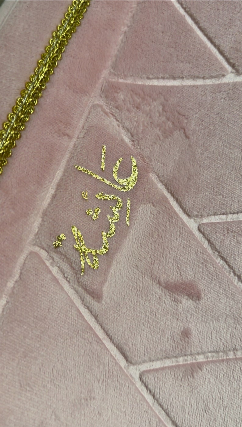 May include: A pink prayer rug with a gold embroidered design. The design features the Arabic word "Allah" in a decorative script.