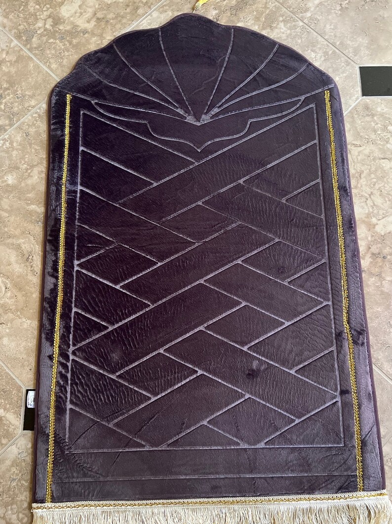 May include: A purple prayer rug with a geometric pattern and gold trim. The rug has a fringed edge and a decorative design at the top.