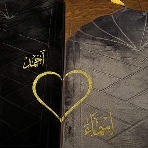 May include: Two black prayer rugs with gold embroidery. The rug on the left has the word "Allah" embroidered in gold. The rug on the right has the word "Insha'Allah" embroidered in gold. A gold heart shape is embroidered between the two rugs.