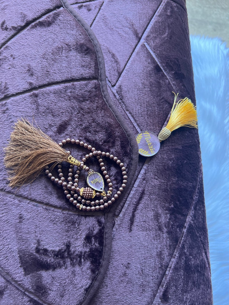 May include: A purple prayer rug with a decorative pattern and a tassel. A brown prayer bead necklace with a tassel and a gold charm with the word "Masjid" is on top of the rug.