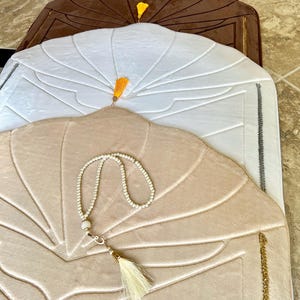 May include: Three prayer rugs with different colored tassels. The top rug is brown, the middle rug is white, and the bottom rug is beige. The beige rug has a white prayer bead necklace and a white tassel.