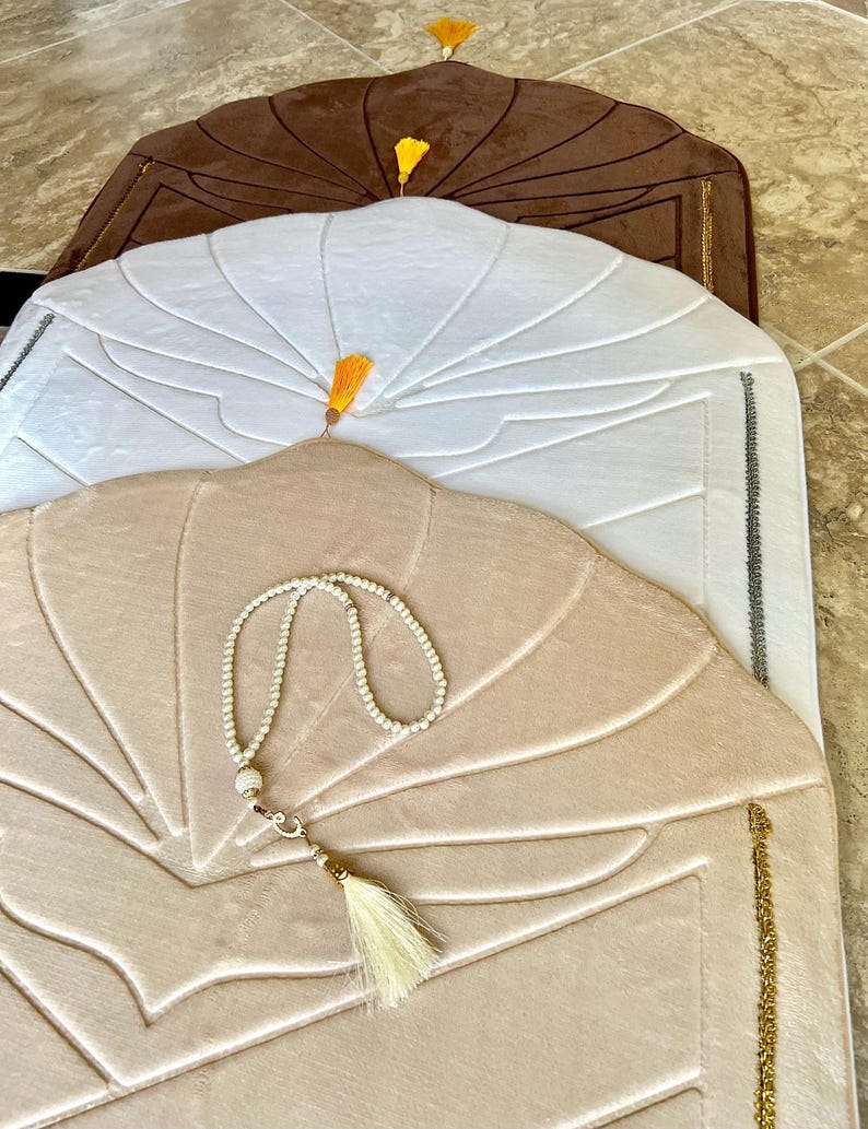 May include: Three prayer rugs with different colored tassels. The top rug is brown, the middle rug is white, and the bottom rug is beige. The beige rug has a white prayer bead necklace and a white tassel.