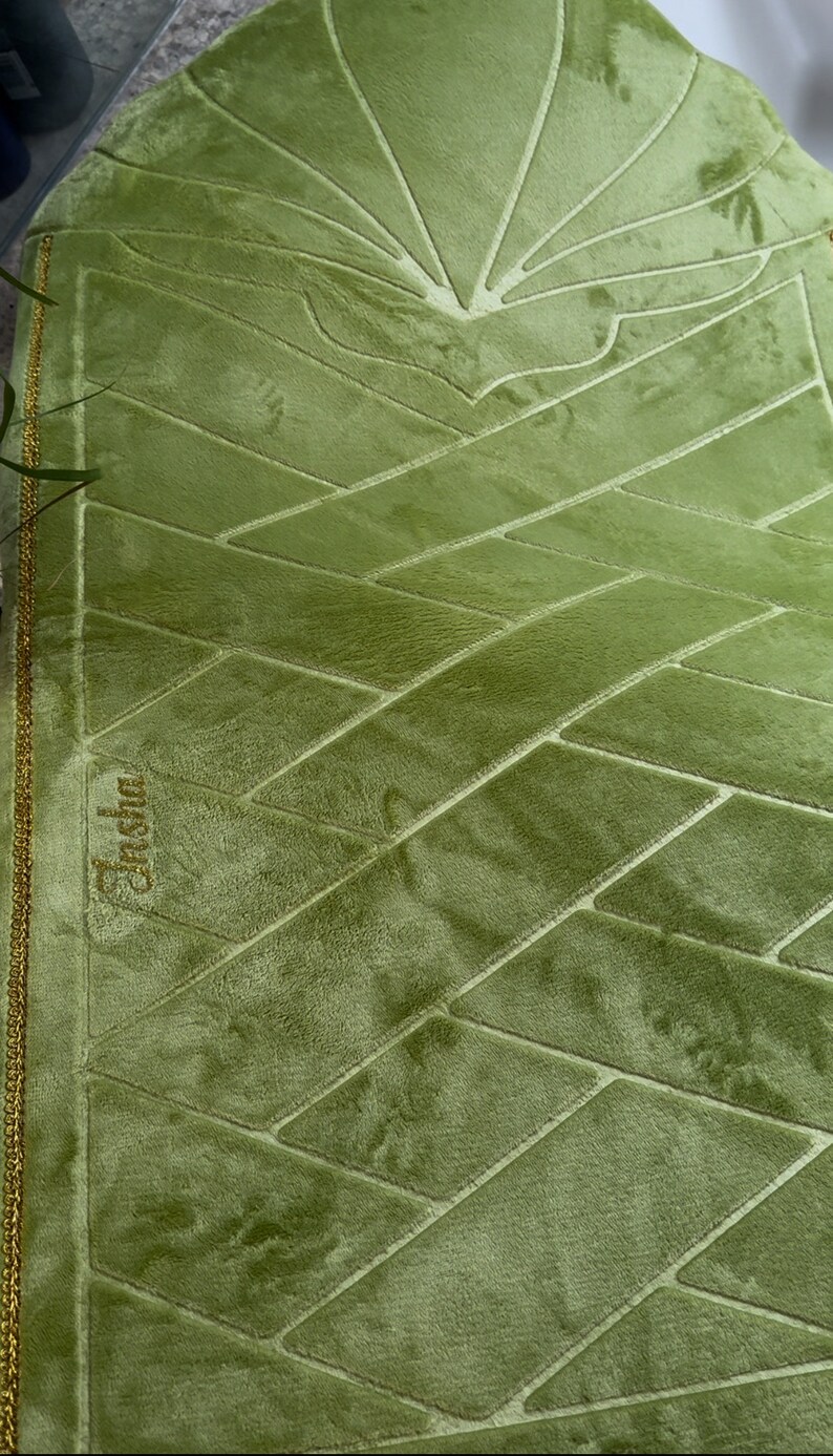 May include: A green prayer rug with a geometric pattern and gold trim. The rug has the word "Jashal" embroidered in gold thread.