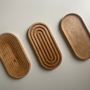 May include: Three wooden oval trays. Two trays have a carved, layered design, while the third is a simple, smooth surface. The trays are made of light-colored wood, showcasing the natural grain. The trays are approximately 12 inches long.