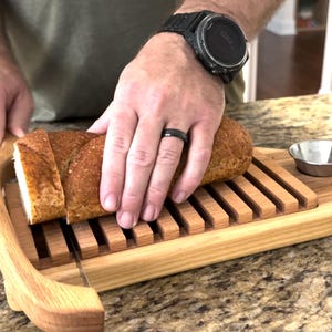May include: Wooden bread slicer with a loaf of bread being sliced. The bread is golden brown, and the slicer has a built-in crumb catcher. A small metal bowl sits on the slicer. The bread is being cut into slices.