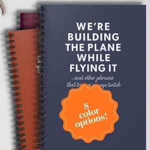 May include: A stack of spiral-bound notebooks in orange, burgundy, and navy blue. The navy notebook has the text "WE'RE BUILDING THE PLANE WHILE FLYING IT" and an orange badge with "8 color options!"