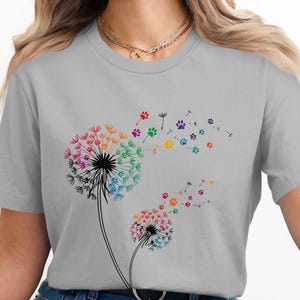 May include: A gray t-shirt with a colorful dandelion design. The dandelion is made up of paw prints in various colors, including red, orange, yellow, green, blue, and purple. The paw prints are arranged in a circle around the center of the dandelion, and the dandelion is blowing in the wind.