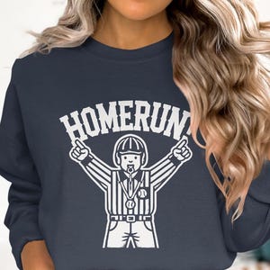 Op de afbeelding: Een marineblauw sweatshirt met een witte afbeelding van een honkbal scheidsrechter die een homerun roept. De tekst "HOMERUN!" staat boven de scheidsrechter.
