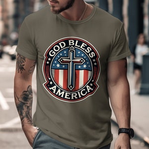 Pode incluir: Uma camiseta verde cáqui com um gráfico circular de estilo vintage que apresenta uma bandeira americana vermelha, branca e azul com uma cruz no centro. O gráfico diz "God Bless America" em uma faixa circular ao redor da bandeira.