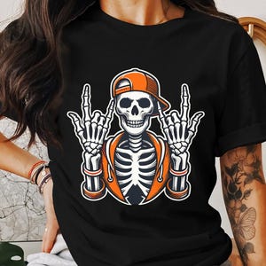 May include: Black t-shirt with a graphic of a skeleton wearing a backwards orange baseball cap and making the 'rock on' hand gesture with both hands.