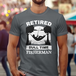 Pode incluir: Uma camiseta cinza escura com o texto "Retired Full Time Fisherman" em branco. O design apresenta uma silhueta de uma pessoa relaxando em uma rede com uma vara de pesca.