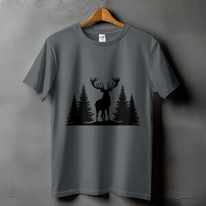 Pode incluir: Uma camiseta cinza com uma silhueta preta de um veado em pé em uma floresta. O veado tem grandes chifres e está rodeado por três pinheiros.
