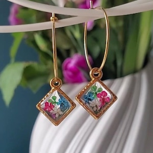 May include: Gold hoop earrings with a square pendant. Each pendant has a diamond-shaped frame filled with colourful dried flowers in pink, blue, and white. The earrings are displayed against a blurred background.