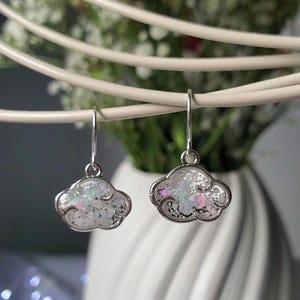May include: Cloud-shaped earrings with silver-coloured frames. The earrings have a white, iridescent centre that reflects multiple colours. The earrings are hanging in front of a white vase with flowers.