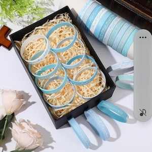 May include: A black gift box filled with light blue and white rubber bracelets with inspirational text. The bracelets are displayed on shredded paper. Additional bracelets and a roll of matching ribbon are also visible.