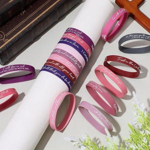 May include: A collection of colorful silicone wristbands with inspirational quotes in white lettering. The bands are in shades of pink, purple, burgundy, and gray. Some bands are stacked on a white cylinder, while others are scattered around. A wooden cross is visible.