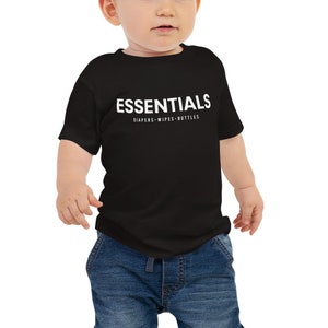 May include: A black baby t-shirt with the word "ESSENTIALS" printed in white across the chest. Below the word "ESSENTIALS" are the words "DIAPERS • WIPES • BOTTLES" in white. The baby is wearing blue jeans.