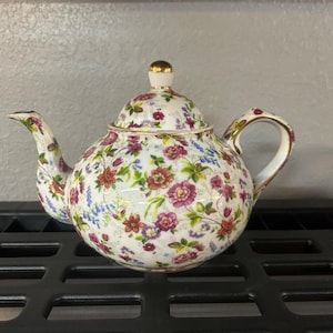 May include: A white ceramic teapot with a floral pattern in shades of red, purple, and green. The teapot has a gold-colored handle, spout, and lid trim. The lid is topped with a gold finial.
