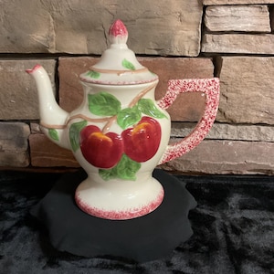May include: Cream-colored ceramic teapot with a lid and handle, decorated with red apples and green leaves. The handle and base have a speckled red design. The spout is curved, and the lid has a pink finial. A decorative kitchenware item.